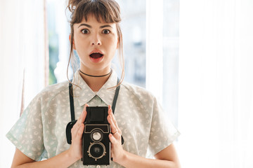Shocked cute young beautiful photographer woman holding camera indoors at home.