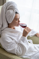 Beautiful young woman in white bath robe lying and drinking wine in hotel room. People, beauty, lifestyle, holidays and relaxation concept