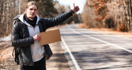 A young man is hitchhiking around the country. The man is trying to catch a passing car for traveling. The man with the backpack went hitchhiking to the south.