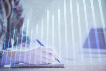 A woman hands writing information about stock market in notepad. Forex chart holograms in front. Concept of research. Double exposure