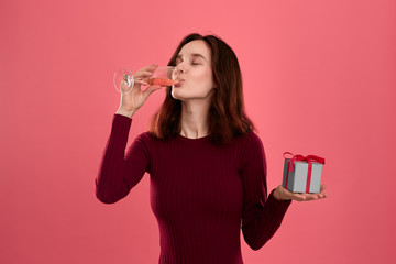 Happy pretty brunette girl holding present box with a ribbon in one hand and drinking champagne wine from a glass standing isolated on a dark pink background. Celebrating special event.