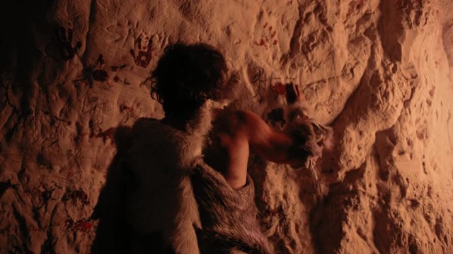 Primitive Prehistoric Neanderthal Wearing Animal Skin Draws Animals and Abstracts on the Walls at Night. Creating First Cave Art with Petroglyphs Rock Paintings Illuminated by Fire. Back View Zoom Out
