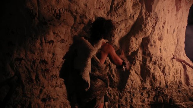 Primitive Prehistoric Neanderthal Wearing Animal Skin Draws Animals and Abstracts on the Walls at Night. Creating First Cave Art with Petroglyphs Rock Paintings Illuminated by Fire. Back View Zoom Out