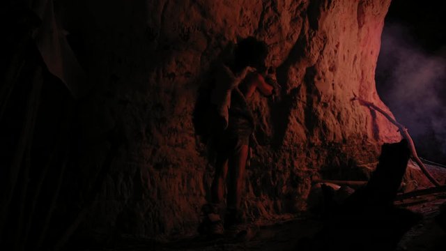 Primitive Prehistoric Neanderthal Wearing Animal Skin Draws Animals and Abstracts on the Walls at Night. Creating First Cave Art with Petroglyphs, Rock Paintings Illuminated by Fire. Back View 