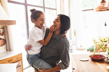 Image of amusing family mother smiling and holding her little daughter in hands at cozy apartment