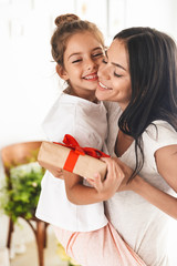 Image of caucasian family brunette mother smiling and giving present box to her little daughter at home