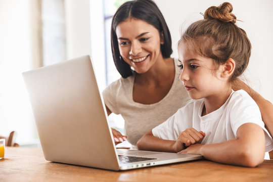 Image of attractive family woman and her little daughter smiling and using laptop computer together in apartment