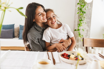 Image of adorable family mother and little daughter hugging while having breakfast at home in morning