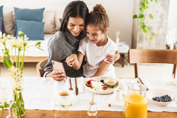 Obraz na płótnie Canvas Image of caucasian family mother and little daughter using cellphone while having breakfast at home in morning