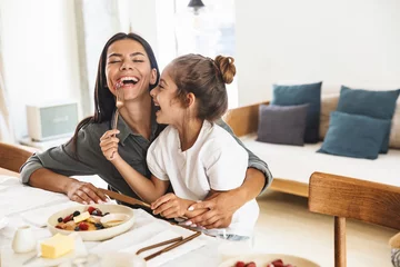Fotobehang Kruidenier Image of cheerful family mother and little daughter smiling and eating together while having breakfast at home in morning  © Drobot Dean