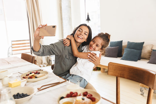 Image of smiling family mother and little daughter taking selfie photo while having breakfast at home in morning