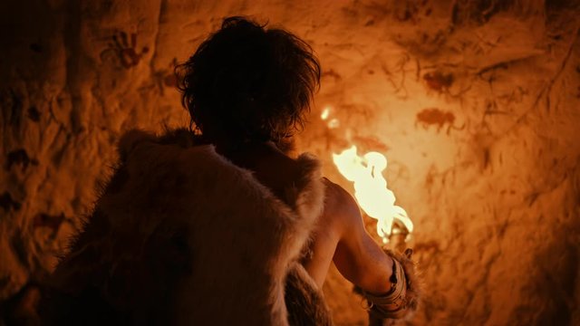 Primeval Caveman Wearing Animal Skin Standing in His Cave At Night, Holding Torch with Fire Looking at Drawings on the Walls at Night. Cave Art with Petroglyphs, Rock Paintings. Back View