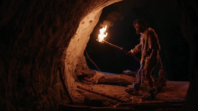 Primeval Caveman Wearing Animal Skin Exploring Cave At Night, Holding Torch with Fire Looking at Drawings on the Walls at Night. Cave Art with Petroglyphs, Rock Paintings. Side View