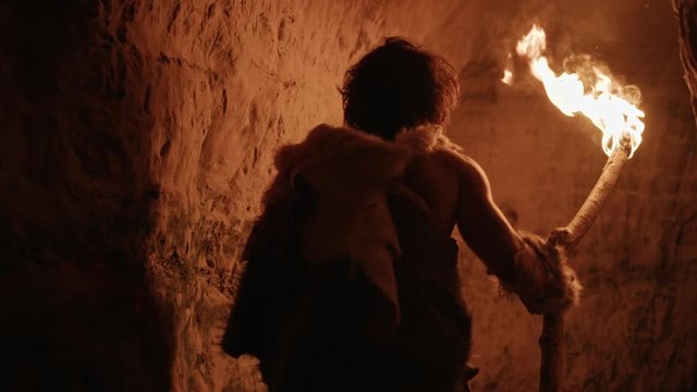 Primeval Caveman Wearing Animal Skin Exploring Cave At Night Holding Torch with Fire Looking at Drawings on the Walls at Night. Neanderthal Searching Safe Place to Spend the Night. Back View Following