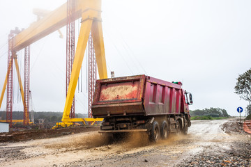 Fototapeta na wymiar Red dirty dump truck moving by dirt road a rainy day near a shipyard