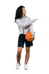A full-length shot of a Young woman playing basketball pointing to the side to present a product over isolated white background