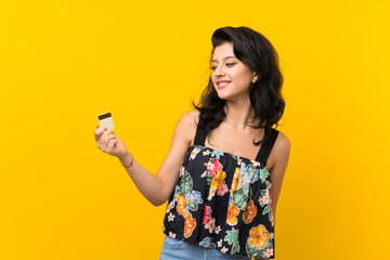 Young woman over isolated yellow background holding a credit card