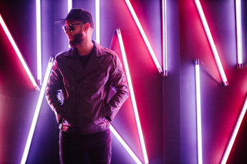 Hipster handsome man on the city streets being illuminated by neon signs. He is wearing leather...