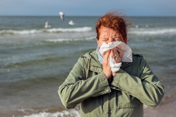 A woman is standing by the sea, using a handkerchief, she has an allergy or severe flu. Concept: Runny nose or protection against infections