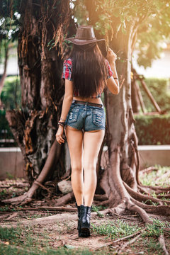 Full-length portrait of sexy young woman in cowboy hat and plaid shirt standing outdoors. Big old tree in the background. Rear view. Vertical shot.