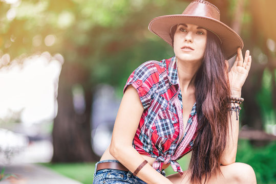 Portrait of sexy woman wearing a cowboy hat looking away pensively while sitting outdoors. Horizontal shot. Countryside lifestyle concept. Selective focus