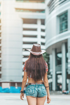 Portrait of young woman in denim shorts and cowboy hat. Country girl came to a big city from a village. Exploring new places. Vertical shot. Rear view. Modern buildings in the background