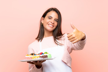 Young girl holding lots of different mini cakes over isolated background with thumbs up because something good has happened