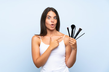 Young girl holding a lot of makeup brush surprised and pointing side