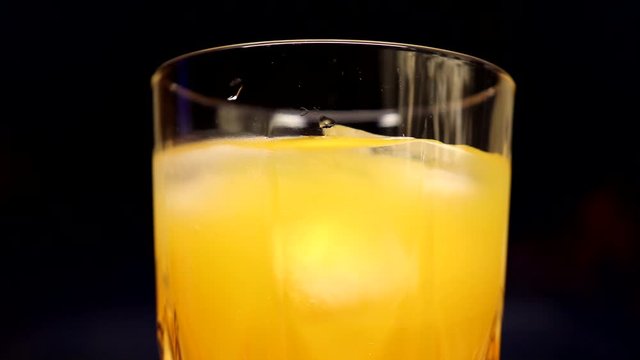 Slowly rotating glass with yellow-orange sweet multifruit juice and ice cubes. Glass with juice on a background of colored smoke.