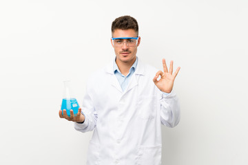 Young scientific holding laboratory flask over isolated background showing an ok sign with fingers