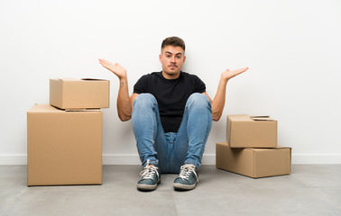 Handsome young man moving in new home among boxes having doubts with confuse face expression