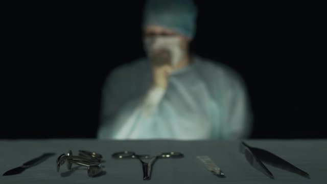 Surgeon thinks about something, reflects before operation or procedure in hospital. Doctor is ponder. Focus on surgical instruments such as scalpel, grasper, forcep, tweezer lie in the foreground