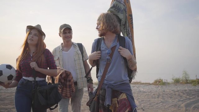 Three young friends came to the beach with guitar and soccer ball on summer evening to relax and have fun