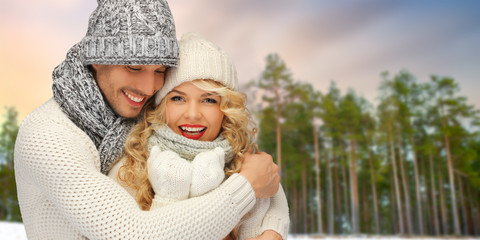 Obraz premium winter, christmas and people concept - happy couple in hats and scarves hugging over forest background