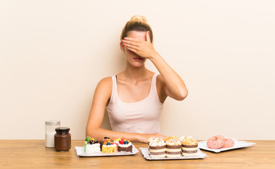 Young woman with lots of different mini cakes in a table covering eyes by hands