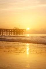 Fototapeta premium Golden sunrise over Atlantic ocean beach. Scenic marine landscape with wooden pier and calm ocean in soft sun light during sunrise at Pawleys Island, South Carolina, USA. Vertical composition.