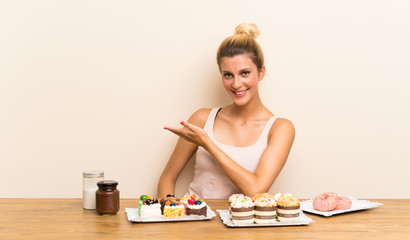 Obraz na płótnie Canvas Young woman with lots of different mini cakes in a table extending hands to the side for inviting to come