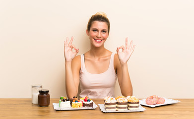 Obraz na płótnie Canvas Young woman with lots of different mini cakes in a table showing an ok sign with fingers