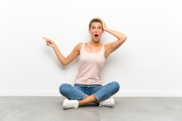Young blonde woman sitting on the floor surprised and pointing finger to the side