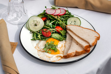 Two sunny side up eggs served with mixed season fresh salad and toasts, on a table outside of cafe or restaurant. Close-up. Tropical vacations concept.