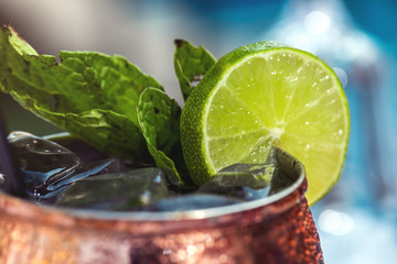 Summer beverage or alcohol drink in bronze glass with slice of lime, mint and straw, and swimming pool as a background. Close up. Tropical summer vacations concept.