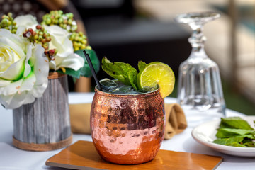 Summer beverage or alcohol drink in bronze glass with slice of lime, mint and straw, served on a wooden plate. Close up. Tropical summer vacations concept.