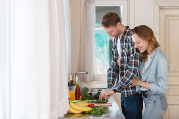 Young happy newlywed couple laughs while cooking vegetable salad in the kitchen of their cozy country house. Concept of happy family life in your own home