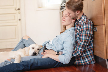 Happy young married couple resting sitting on the floor near the kitchen set hugging their beloved dog. Holiday concept during the weekend