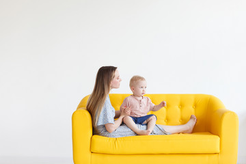 Charming young mother in home dress is sitting on a yellow sofa and is holding a her cute little son in the arms. Concept of care for small children and decree. Advertising space