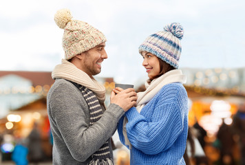 people, winter holidays and love concept - happy romantic couple in knitted hats and scarves holding one cup over christmas market in old town of tallinn city background