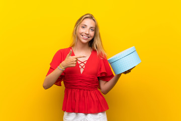 Blonde young woman over isolated yellow background holding gift box