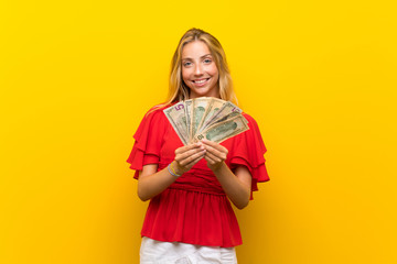 Blonde young woman over isolated yellow background taking a lot of money