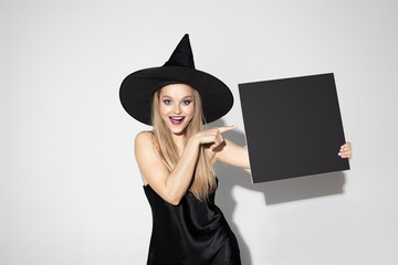 Young blonde woman in black hat and costume on white background. Attractive caucasian female model. Halloween, black friday, cyber monday, sales, autumn concept. Copyspace. Holding black copyspace.