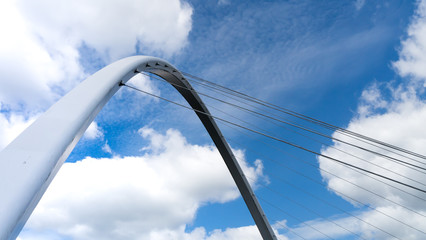 The Gateshead Millennium Bridge that spans the River Tyne connecting Gateshead to Newcastle. Image showing the arch and steel cables of the bridge contrasting against a blue cloudy sky in summer.